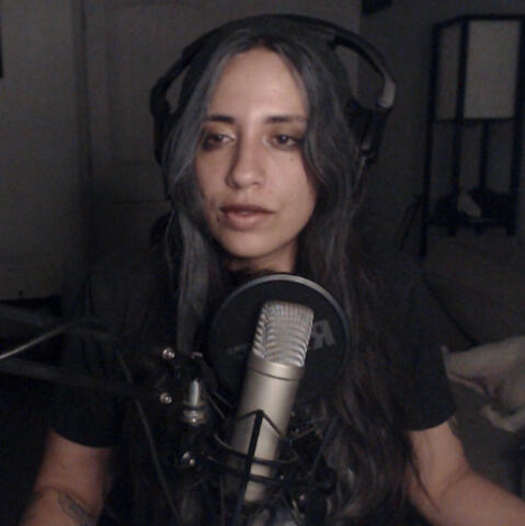 A woman with long dark hair and green slouchy beanie posing with a Peace sign in front of a mic. Eli
